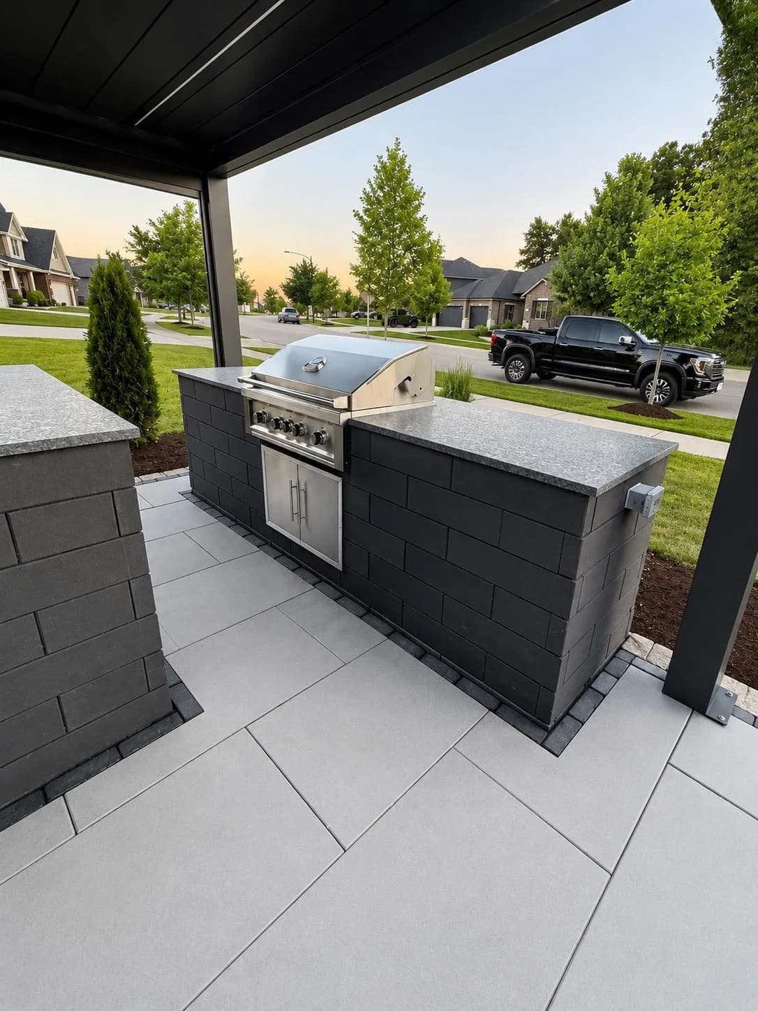 Outdoor kitchen with a built-in stainless steel gas grill set into a charcoal paver island with granite countertop, under a black pergola, overlooking a suburban street at sunset.