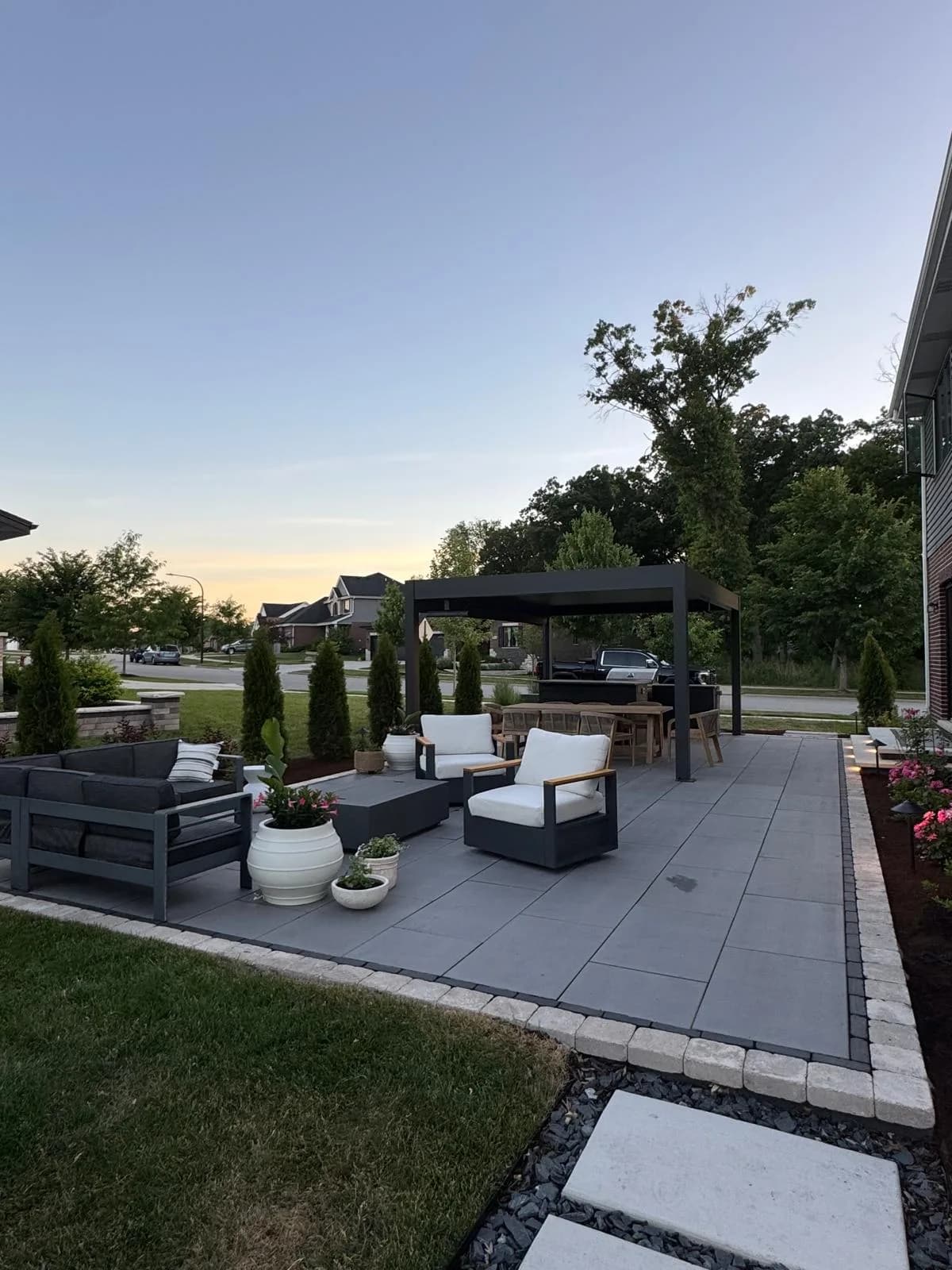 Modern backyard paver patio at dusk with a black aluminum pergola, charcoal sectional sofa, fire table, and landscaped garden beds beside a brick-and-siding home.