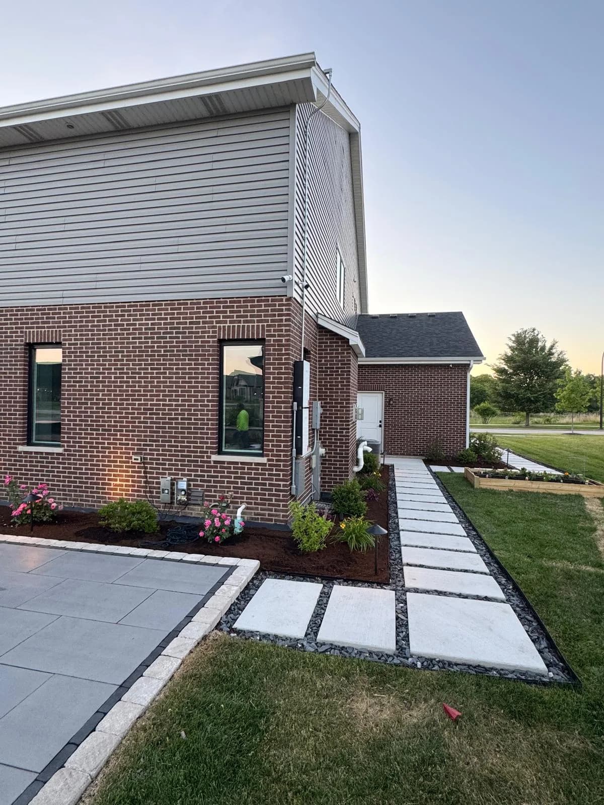 Large-format concrete paver walkway with black gravel and dark border edging running alongside a brick home, framed by fresh mulch beds, flowers, and a raised garden planter.