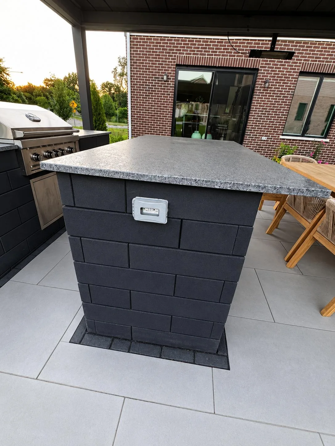 Custom outdoor kitchen island clad in charcoal paver veneer with a polished granite top and built-in electrical outlet, beside a stainless steel grill and teak dining set.