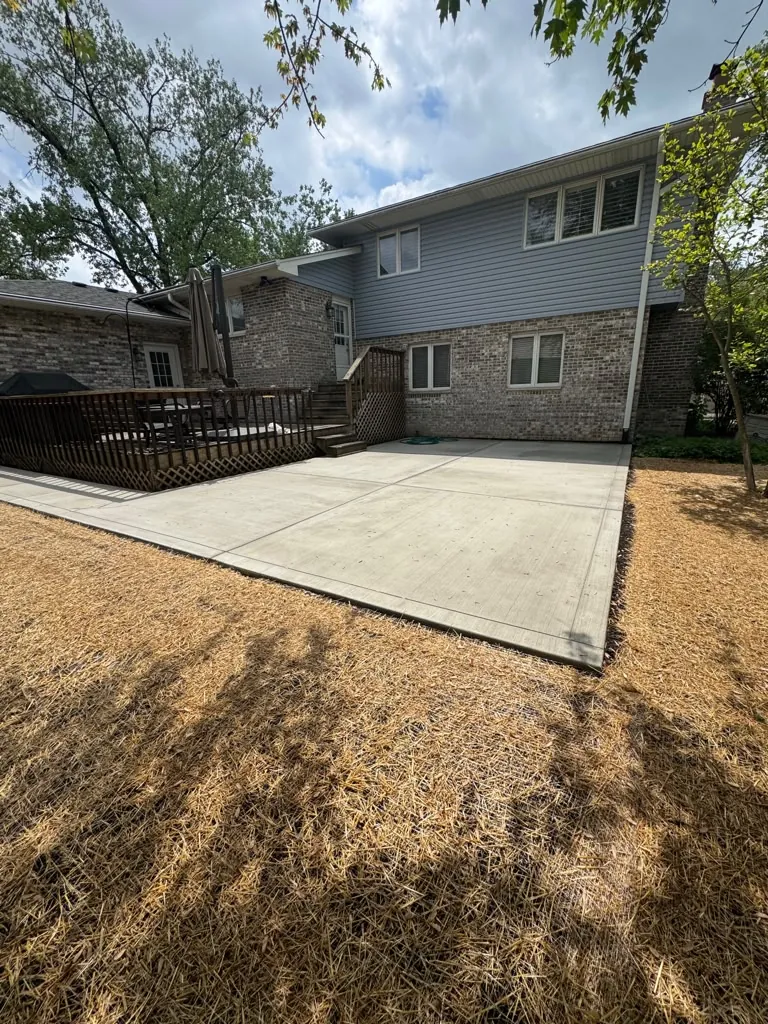 Large poured concrete patio behind a brick and blue-sided house with a freshly seeded straw-covered yard in Chicago's Southwest Suburbs