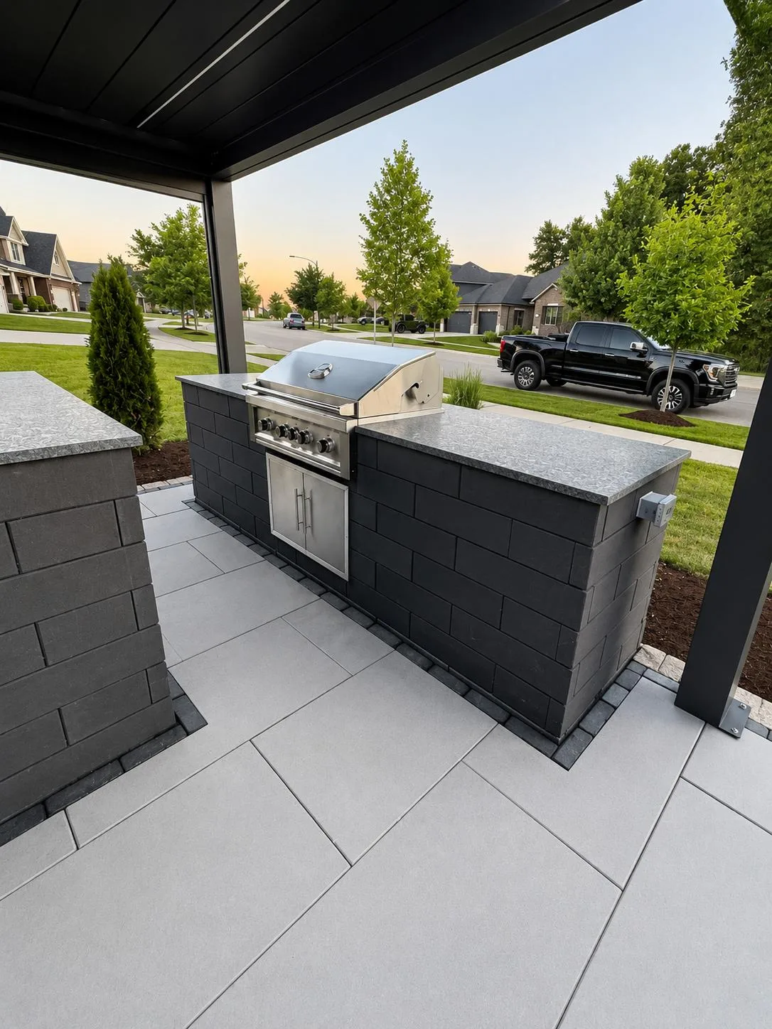 Outdoor kitchen with a built-in stainless steel gas grill set into a charcoal paver island with granite countertop, under a black pergola, overlooking a suburban street at sunset.