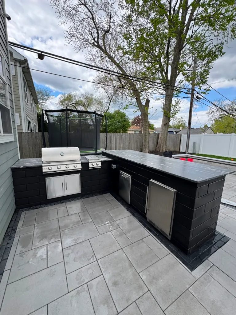 L-shaped black block outdoor kitchen with a stainless grill, side burner, fridge, doors, and dark stone countertop on a gray paver patio in Chicago's Southwest Suburbs