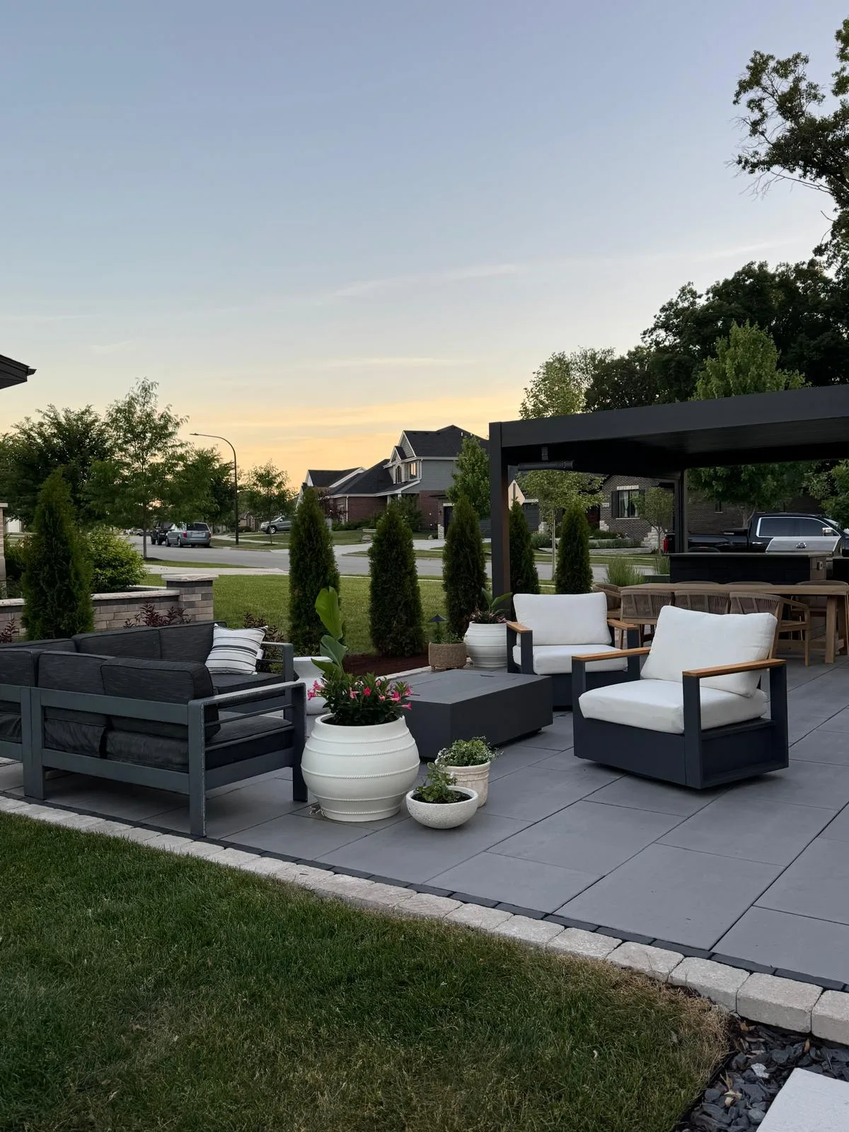 Golden-hour paver patio with a charcoal sofa and white-cushioned lounge chairs, large white planters with greenery, a fire table, and a pergola-covered dining area in the background.