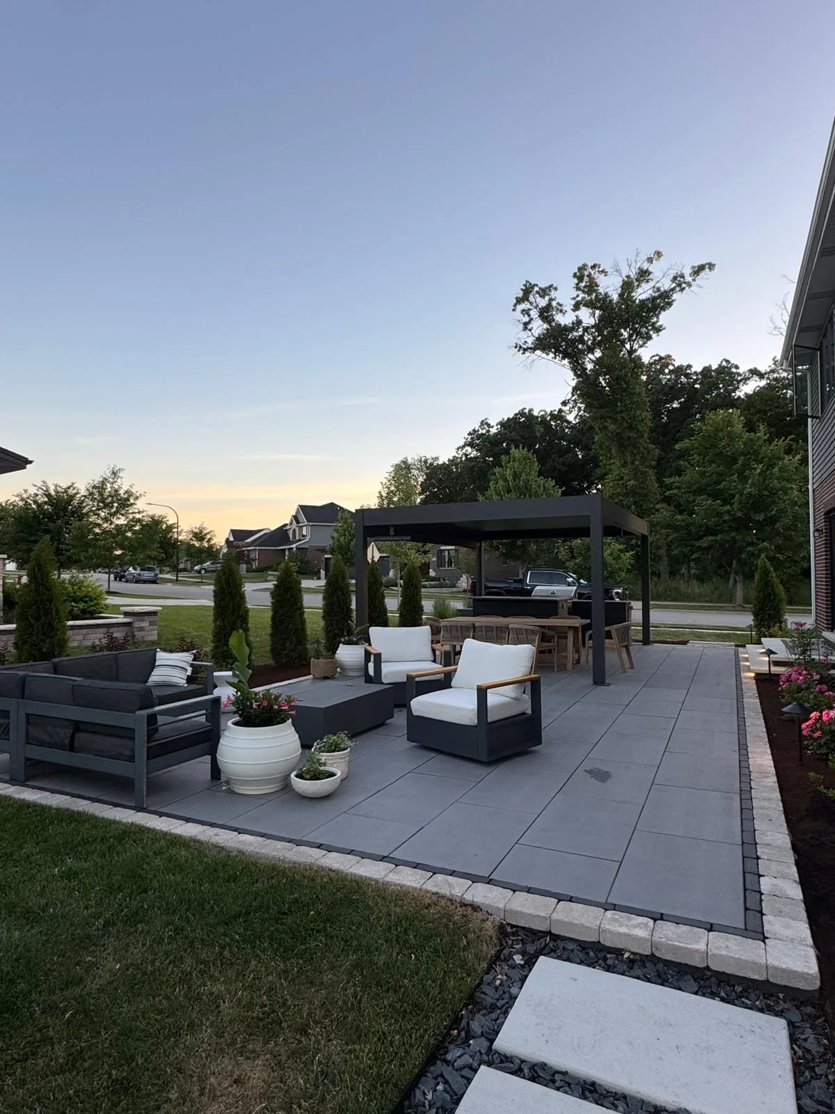 Modern backyard paver patio at dusk with a black aluminum pergola, charcoal sectional sofa, fire table, and landscaped garden beds beside a brick-and-siding home.