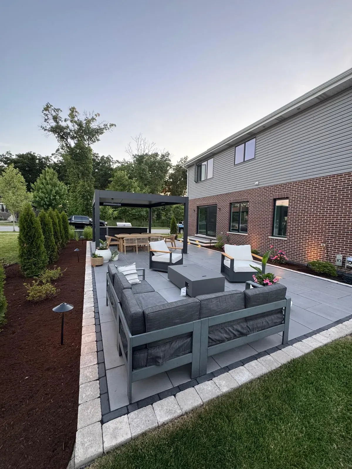 Wide view of a large-format grey paver patio with charcoal lounge furniture, white cushioned chairs, a black pergola, and a paver stepping-stone path leading across the lawn.
