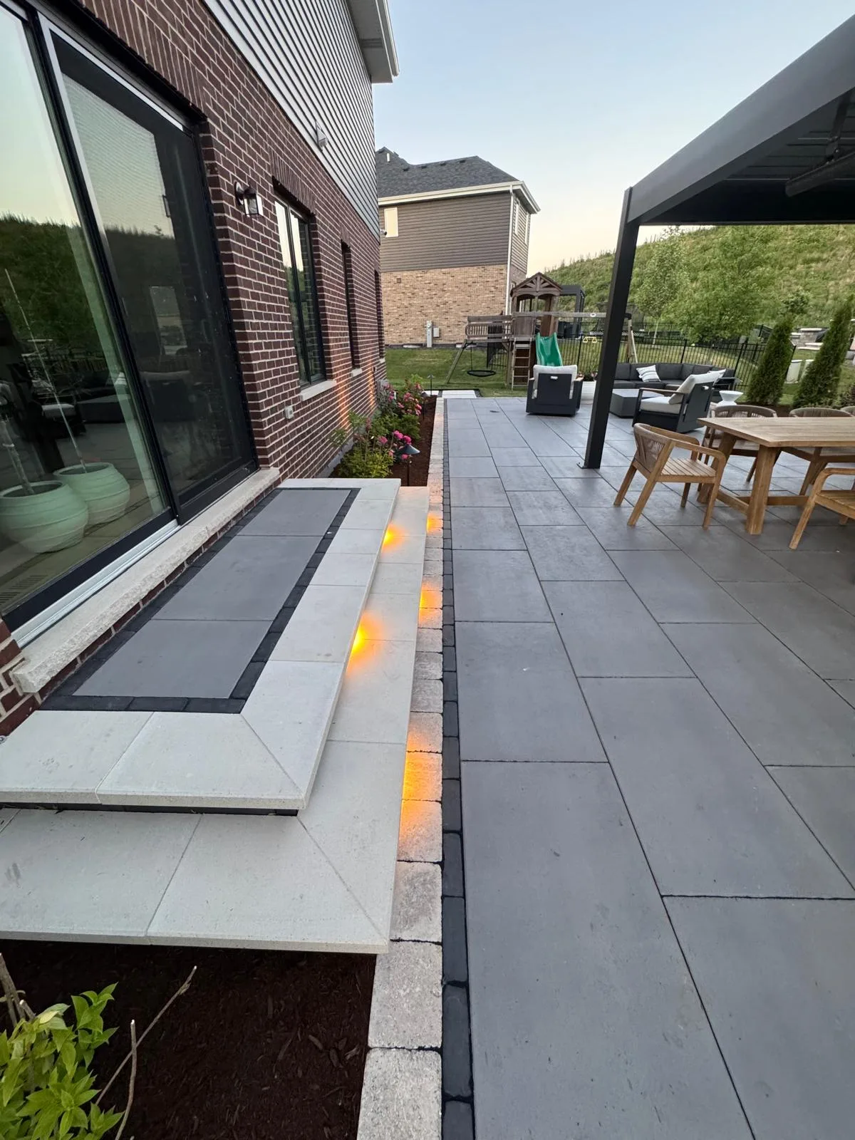 Evening view down a paver patio showing a raised seating ledge along the brick home wall with warm integrated LED step lighting glowing beneath the cap stones.