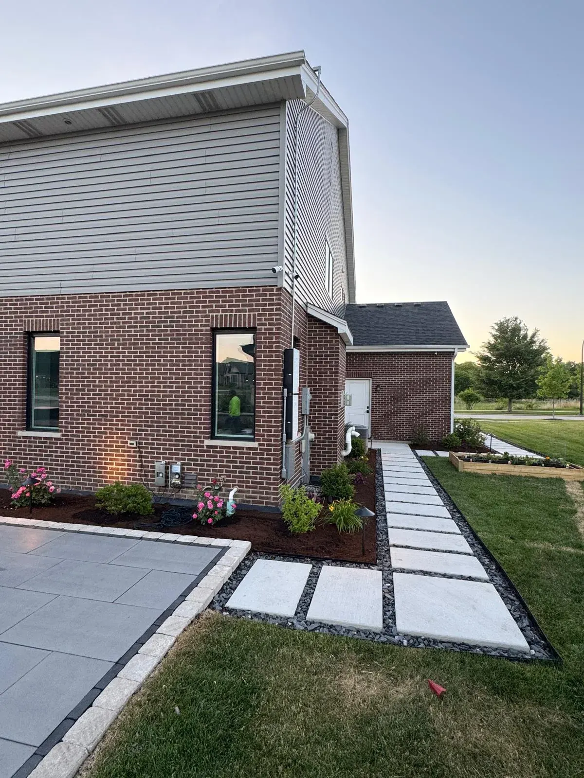 Large-format concrete paver walkway with black gravel and dark border edging running alongside a brick home, framed by fresh mulch beds, flowers, and a raised garden planter.