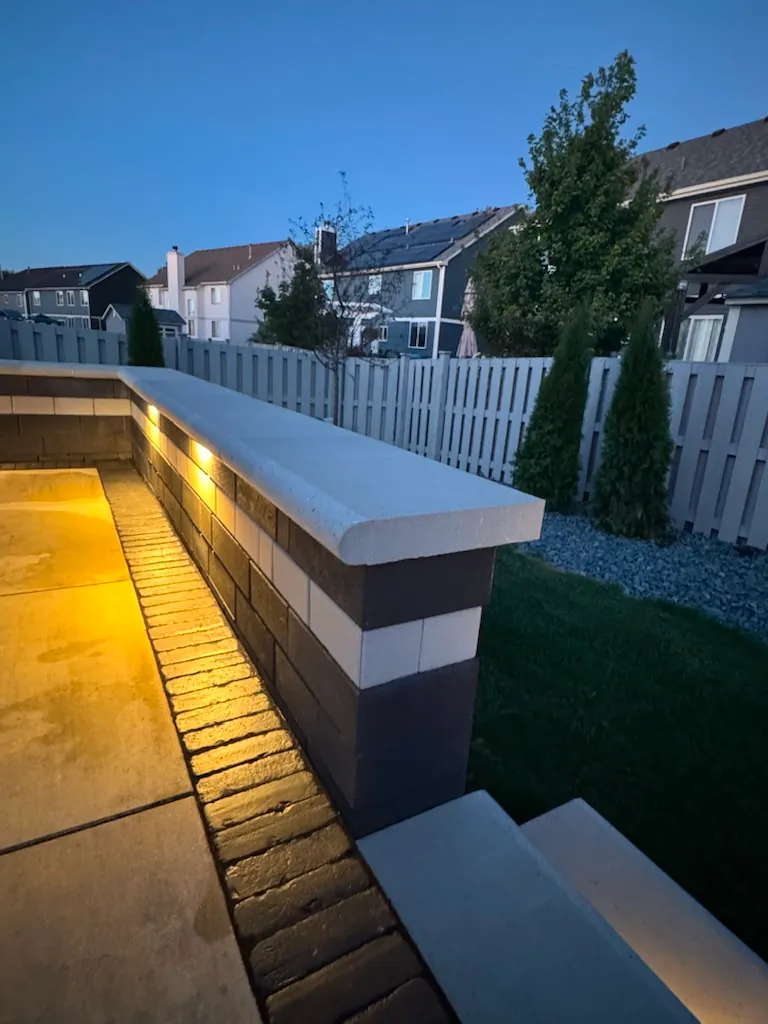 Long two-tone block seat wall with smooth cap stone and warm under-cap LED lighting at dusk beside a concrete patio in Chicago's Southwest Suburbs