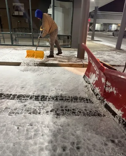 Crew member shoveling a snow-covered commercial sidewalk at night beside a red plow blade in Chicago's Southwest Suburbs