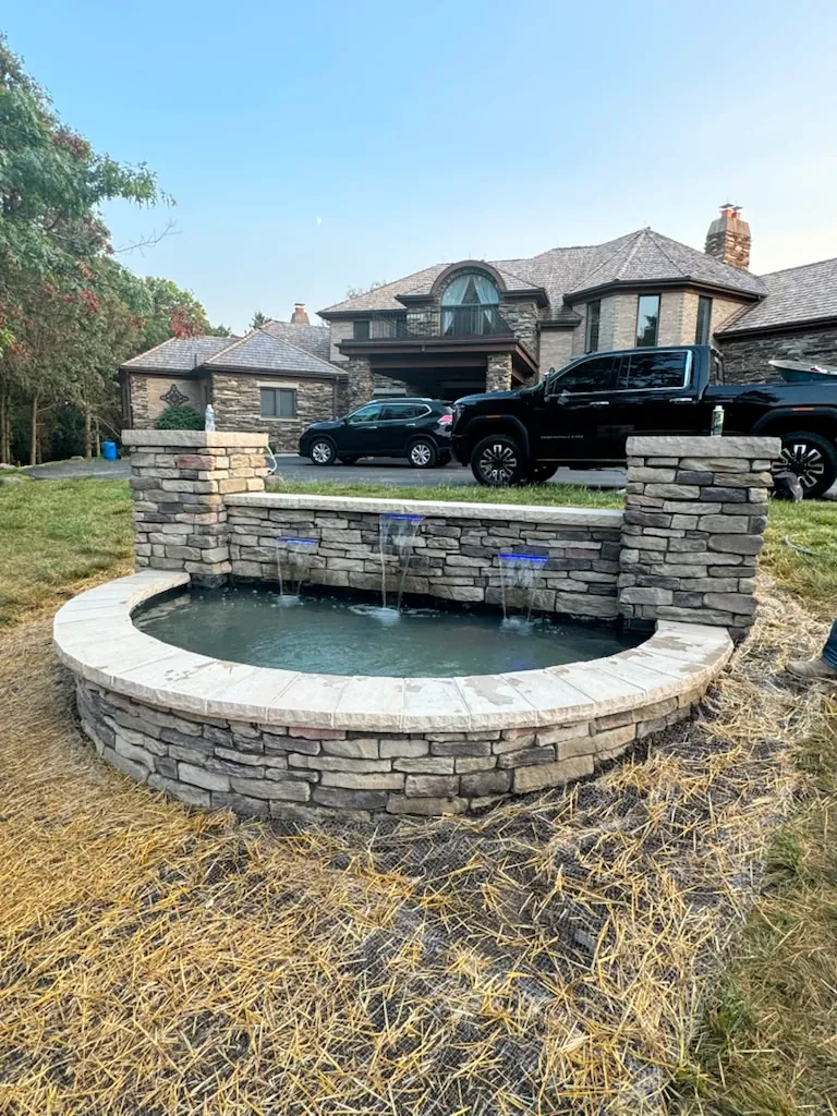Semicircular stacked-stone raised water feature with scuppers and blue lighting in front of a large home in Chicago's Southwest Suburbs